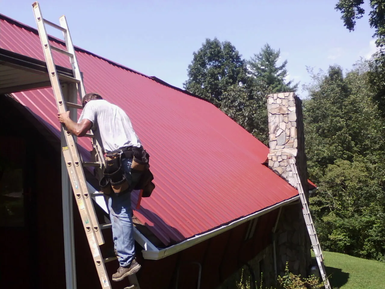 Roofer on a ladder beside a red metal roof and stone chimney — Soffit & Fascia Repair by Roofing Brothers in Bandera